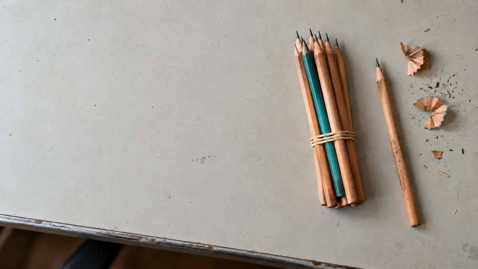 Un faisceau de crayons tenu par un élastique, dont un crayon teal, à côté d’un crayon seul sur un bureau gris.